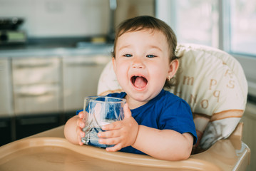 A little girl in the kitchen in the afternoon in a high chair drinking water from a glass glass