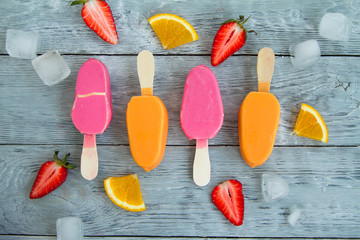 four pink and yellow ice creams on a stick on a gray wooden background next to juicy pieces of...