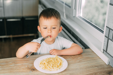 little boy eats pasta in the form of a spiral in the afternoon in the kitchen on their own