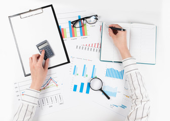 The hands of a woman count on a calculator and write notes. Top view of business woman's hands on white desk with graphs. Finance and business concept.