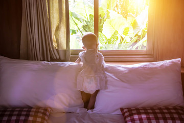A little girl in a white dress is standing on the bed. Looks out the window. There is tropical vegetation. A infant child of eight months leaned on the window sill.