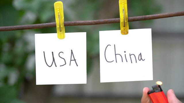 Inscriptions on white stickers on yellow clothespins on dark background. Papers with words USA on left and China on right hanging on metal rod. Man&rsquo;s hand use red lighter ignites the word on right.