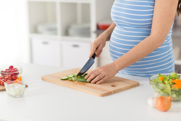 Obraz na płótnie Canvas pregnancy, cooking food and healthy eating concept - close up of pregnant woman making vegetable salad and chopping cucumber by kitchen knife on cutting board at home kitchen
