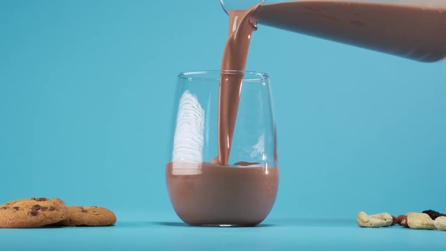 Hand holding jug pours chocolate milk in to glass standing on blue background.