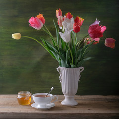 A white cup of tea, a jar of honey and a bouquet of tulips in a vase on the table