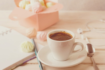 Morning coffee with a notebook and a pencil, with a box filled with small meringues, headphones on a wooden light background.