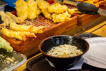 A bowl of udon on a tray and several varieties of tempura side dishes on the counter