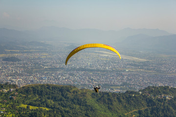 paraglider on a yellow parachute flies against the background of the city in a green mountain valley in the fog, aerial view