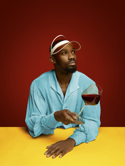 Young man as a medieval grandee or nobleman on red studio background. Holding a glass of red wine. Portrait in retro costume and a cap. Human emotions, comparison of eras, facial expressions concept.
