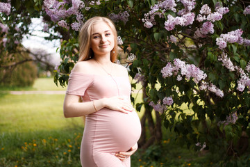 Fototapeta na wymiar pregnant blonde girl in pink dress with bare shoulders holding hands belly with long hair on the background of lilacs and trees with a big belly on the eighth month of pregnancy smiling