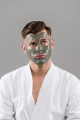 front view of sad young man in bathrobe with clay mask isolated on grey