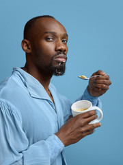 Young man as a medieval grandee on blue background. Portrait of male african-american model in retro shirt. Eating icecream in a cap. Human emotions, comparison of eras, facial expressions concept.