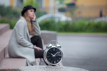 a girl sits on the steps and waits, a blurred silhouette, a clock in the foreground in focus, being late for a date, a long wait