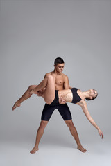 Obraz premium The couple of a young modern ballet dancers in black suits are posing over a gray studio background.