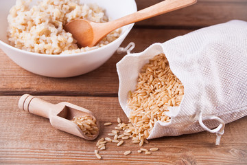Dry rice in a bag and cooked brown rice in white bowl on the wooden background.