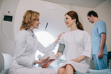 smiling doctor measuring patients blood pressure with tonometer while assistant adjusting ct scanner