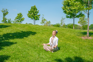 Naklejka na ściany i meble Beautiful boho style woman with accessories is meditating sitting in Lotus pose on grass in park.