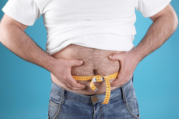 Overweight man with large belly and measuring tape on color background, closeup