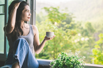 beautiful Asian woman sitting next to the window drinking coffee and relaxing with beautiful nature...