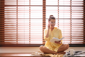 Young woman reading book near window with blinds at home. Space for text - Powered by Adobe