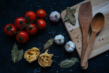 Fresh raw tomatoes, garlic, pasta and wooden spatula and a spoon on a wooden cutting board isolated on black table background. Top view. Pasta ingredients