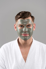 front view of smiling young man with clay mask in white bathrobe isolated on grey