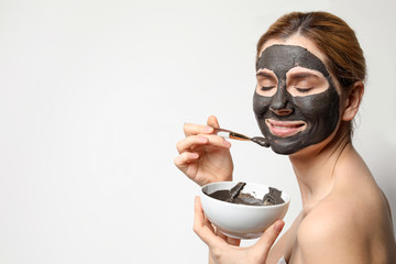 Beautiful woman applying black mask onto face against light background. Space for text