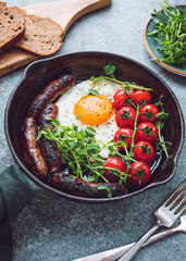 Breakfast time, fried egg with sausages and cherry tomatoes in a black iron pan, served microgreens.