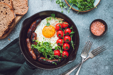 Breakfast time, fried egg with sausages and cherry tomatoes in a black iron pan, served microgreens. Top view, flat lay.