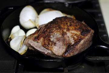 Sirloin steak frying in a cast iron pan with onions on the stove top.