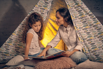 Mother and daughter are sitting in a teepee tent with some pillows and reading stories. Happy...
