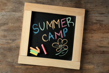 Small blackboard with text SUMMER CAMP, drawing and chalk sticks on wooden background, top view