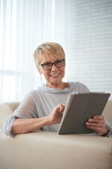 Portrait of mature woman with short hair wearing eyeglasses working online on digital tablet while sitting on sofa at home