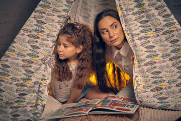 Mother and daughter are sitting in a teepee tent with some pillows and reading stories. Happy...
