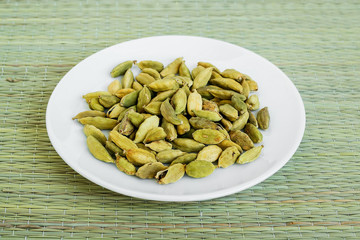 A lot of dry cardamom seeds on a white saucer on a green table mat made of natural plant fibers. Natural food spices and seasonings.