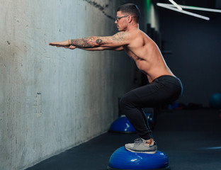 Young strong man in gym doing squats on a fitness hemisphere