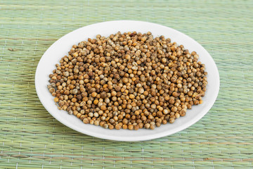 A lot of coriander seeds on a white saucer on a green table mat made of natural plant fibers. Natural food spices and seasonings.
