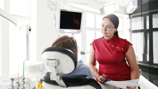 A young girl at a consultation with a dentist woman in glasses is sitting on a chair in a stamotology office. The conversation of the doctor and the patient