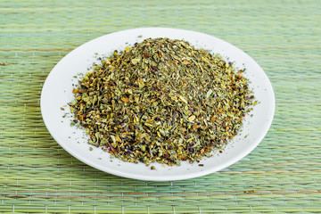 Fototapeta na wymiar A heap of dry basil flakes on a white saucer on a green table mat made of natural plant fibers. Natural food spices and seasonings.