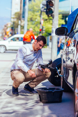 A strong businessman is bending his knee while trying to change a flattie on his car with his lug wrench.