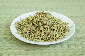 A heap of dry rosemary on a white saucer on a green table mat made of natural plant fibers. Natural food spices and seasonings.