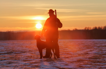 A hunter in a hat with a gun and a German shorthair pointer dog breed stands on a meadow covered with snow. Majestic winter scenery. Amazing sunrise.