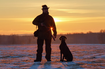 A hunter in a hat with a gun and a German shorthair pointer dog breed stands on a meadow covered...