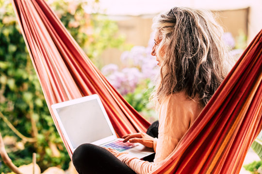 Alternative style caucasian people long grey white hair adult woman enjoy technology outdoor at home sit down on a red hammock writing on a modern laptop computer - garden in background