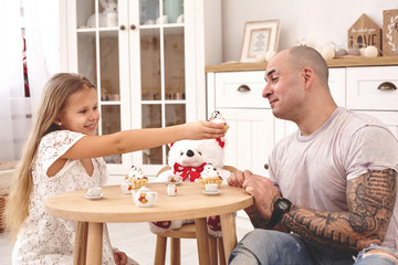 Adorable daughter wearing a white dress whith her loving father. They are drinking tea from a toy dishes in a modern kid's room whith a wooden furniture. Happy family.