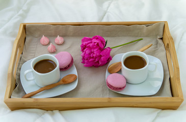 Breakfast in bed for two. Wooden tray with coffee, macaroons and Bizet. Decoration pink peony. Beautiful natural light from the window.