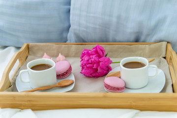 Breakfast in bed for two. Wooden tray with coffee, macaroons and Bizet. Decoration pink peony. Beautiful natural light from the window.