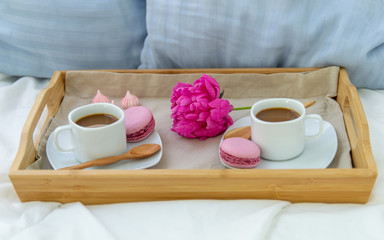 Breakfast in bed for two. Wooden tray with coffee, macaroons and Bizet. Decoration pink peony. Beautiful natural light from the window.