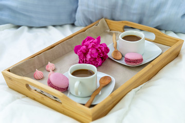 Breakfast in bed for two. Wooden tray with coffee, macaroons and Bizet. Decoration pink peony. Beautiful natural light from the window.