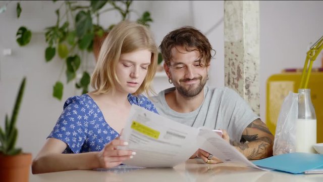 Stressed young blonde girl looking at important documents with sad boyfriend on morning, solving domestic paper problems together sitting at cosy flat with green plant and bottle of milk on background